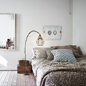 Cozy bedroom interior with neutral tones. Features a bed with textured bedding, decorative pillows, and a metal floor lamp with a woven shade.