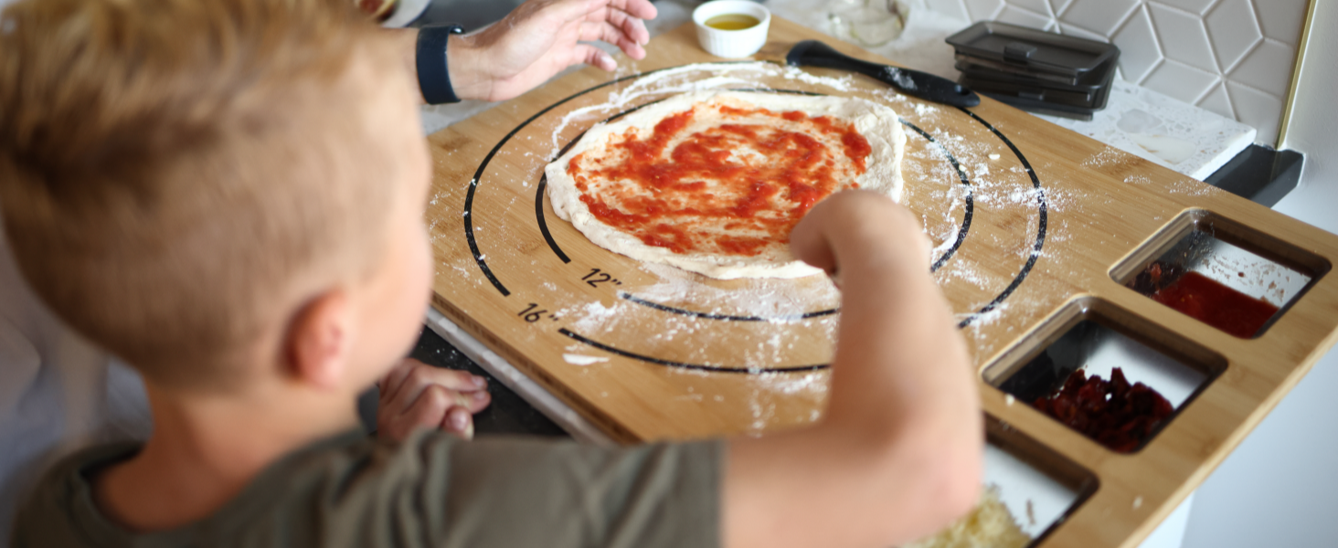 Wooden pizza board or cutting surface with tomato sauce being spread in circular motion, surrounded by prep ingredients and toppings.