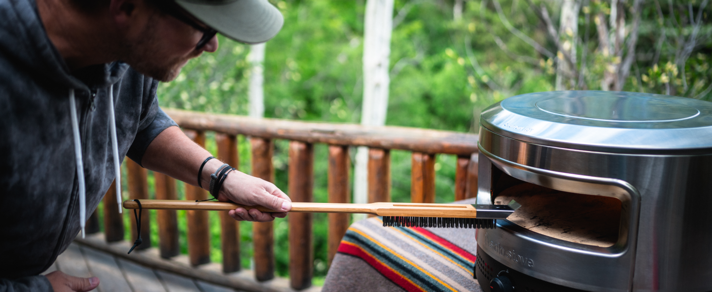Person inserting food into a stainless steel outdoor pizza oven on a wooden deck. The oven has a cylindrical shape with a front opening.