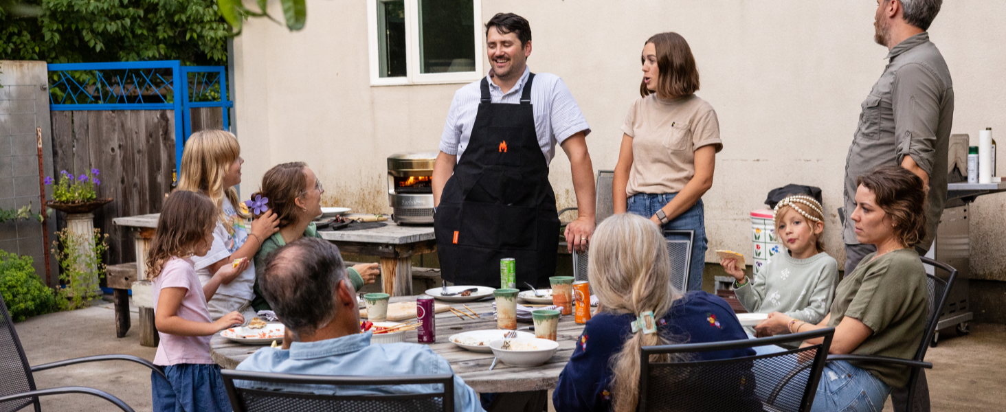 Outdoor dining scene with multiple people seated around a table. Man in apron standing, suggesting a barbecue or casual gathering setting.