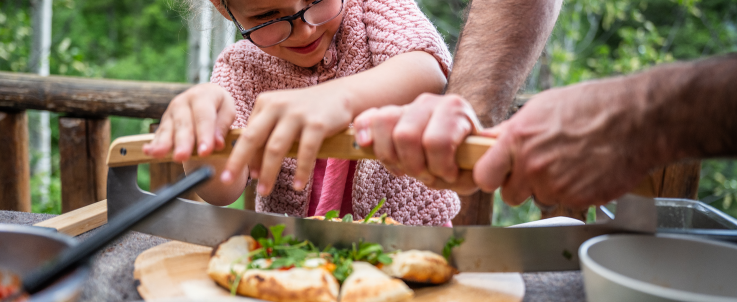 Large serrated knife cutting bread on wooden board. Multiple hands visible, demonstrating collaborative food preparation in outdoor setting.